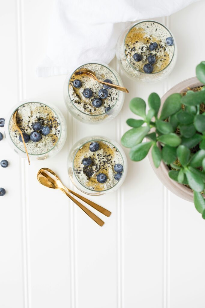 a healthy breakfast scene with 4 jars of chia pudding with honey and blueberries arranged on a board near a green plant