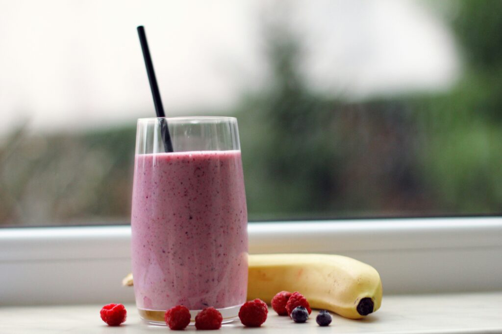 pinkish purple smoothie in a clear glass sitting on a windowsill with a black straw, a banana, raspberries, and blueberries around the bottom of the glass